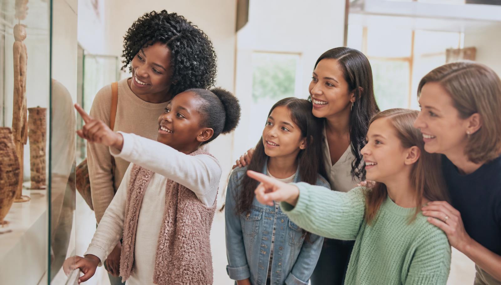 Mothers and daughters of different ages and backgrounds smiling together as they point at a museum exhibit